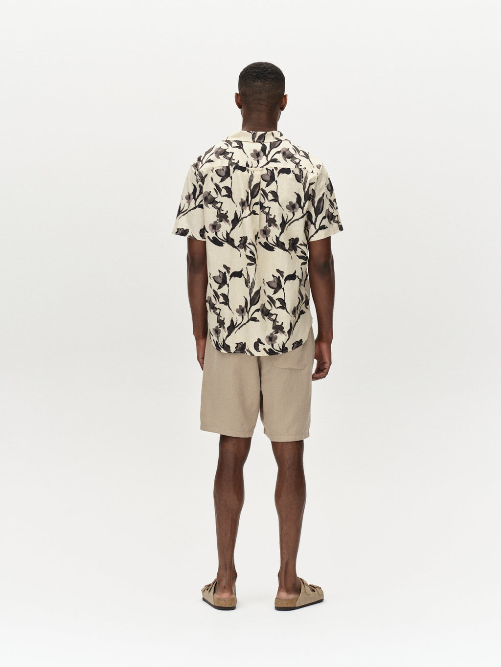 A man stands against a white background, wearing the Alfa Lino Sand Plaza beige shirt featuring a dark leafy pattern and premium comfort shorts. Sandals complete his look. All from Gabba.