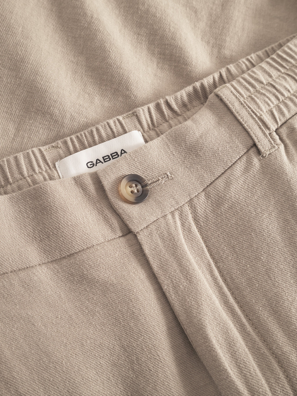 Close-up of sand-colored Alfa Lino pants from Gabba, showcasing a button closure with an adjacent clasp and elastic waistband for superior comfort. A white label displaying Gabba is above the waistband.