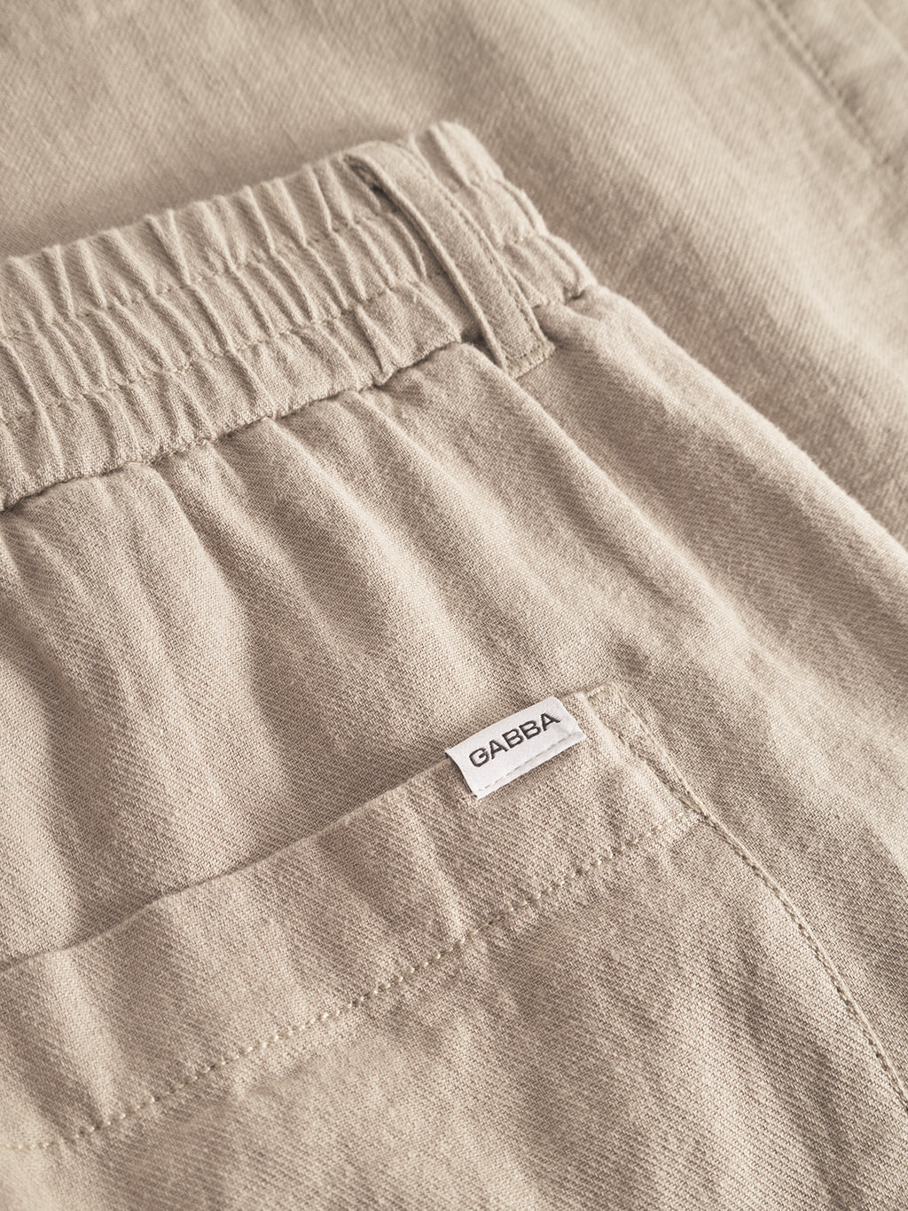 A close-up of the Alfa Lino - Sand Plaza pants highlights their superior comfort, featuring an elastic waistband and a small rear pocket with a Gabba label. The beige textured fabric looks soft, ideal for casual wear.
