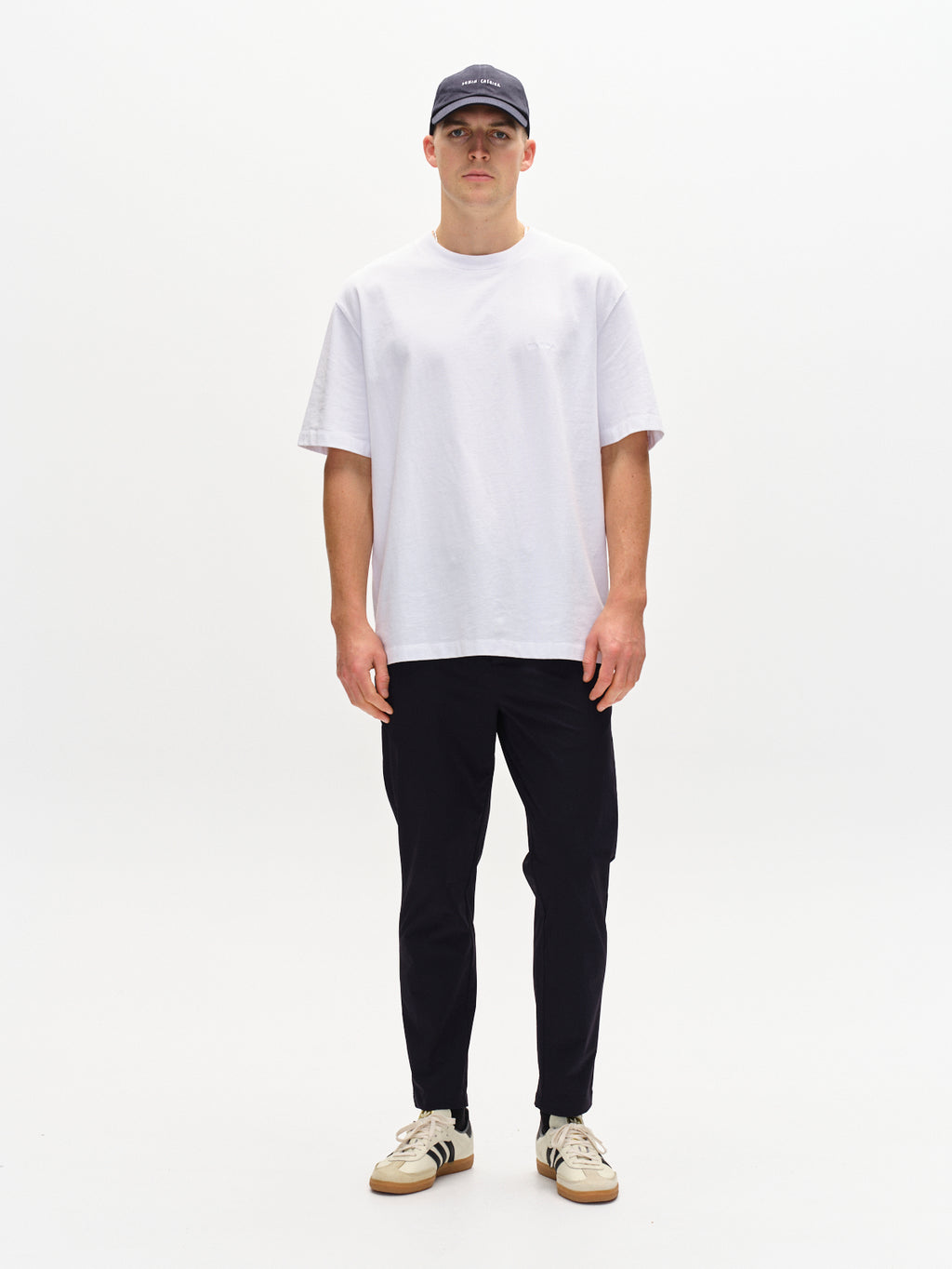 Facing forward against a plain white background, they wear a black cap, a plain white T-shirt, relaxed straight-fit black pants with an elastic waist, and complete the look with Gabbas Monza Tech - Navy sneakers featuring black stripes.