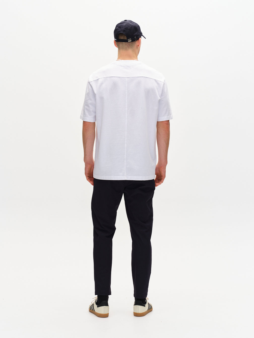 A person showcases Gabbas Monza Tech navy relaxed straight fit pants and a white T-shirt. Theyre wearing a black cap and white sneakers with black stripes, effortlessly contrasting the plain white background.