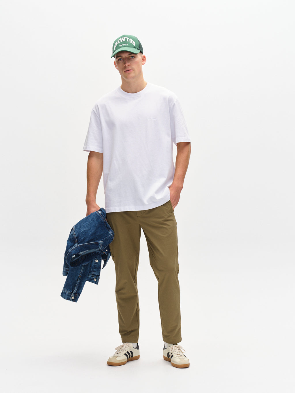 A person wearing a green cap, casual white T-shirt, Monza Tech - Army olive pants with an elastic waist by Gabba, and sneakers stands holding a denim jacket. The plain white background enhances the minimalist look with relaxed straight fit vibes.