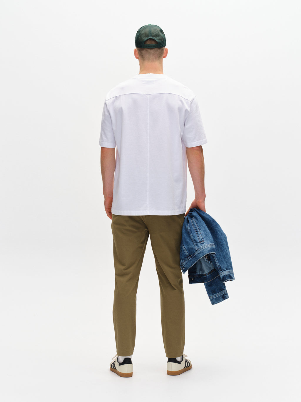 A person stands with their back to the camera, wearing Gabbas Monza Tech - Army olive pants in a relaxed straight fit, a white T-shirt, sneakers, and a green cap. They hold a denim jacket while standing against a plain white background.