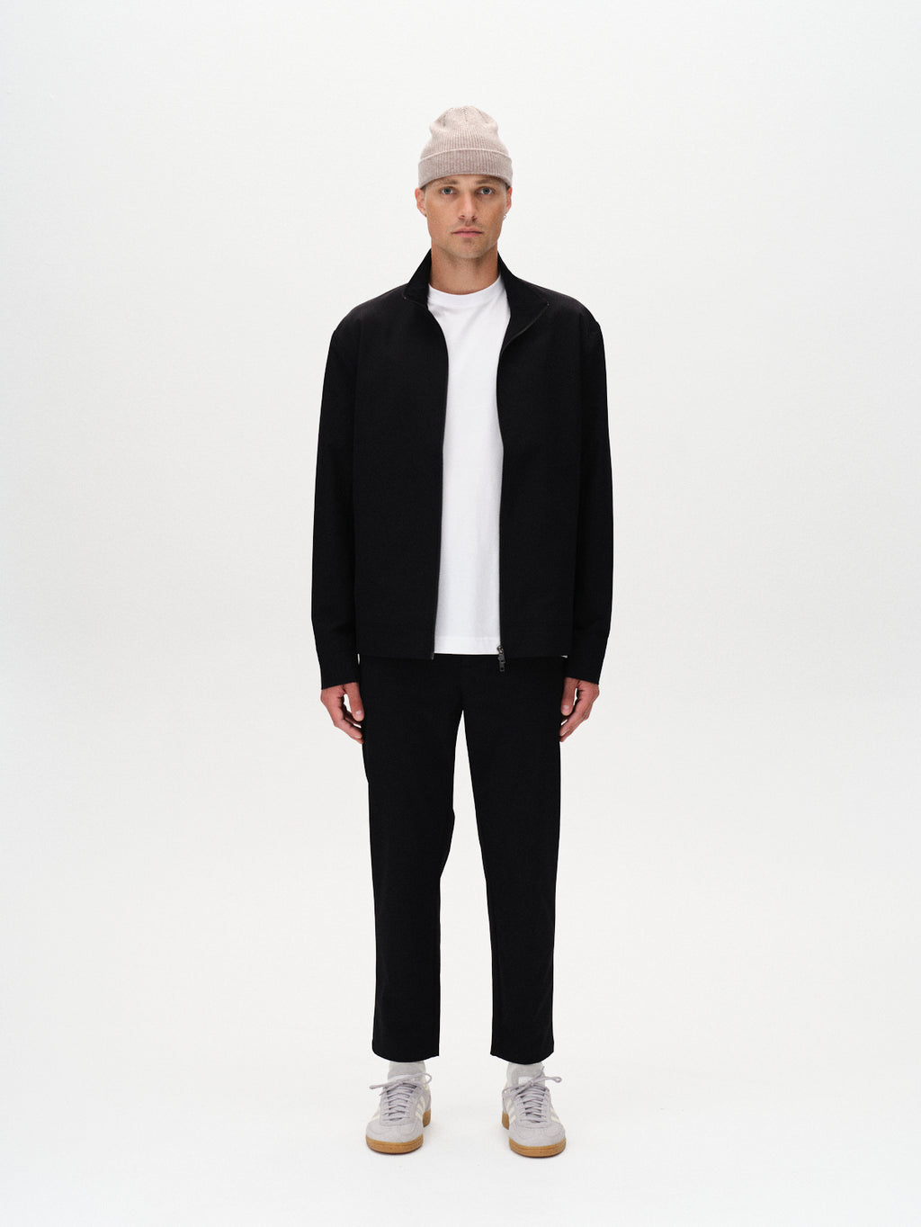 A person stands facing forward against a white background, wearing Gabba’s Monza Tech - Black zip-up jacket, a beige beanie, a white t-shirt, black relaxed straight fit pants with an elastic waist, and white sneakers.