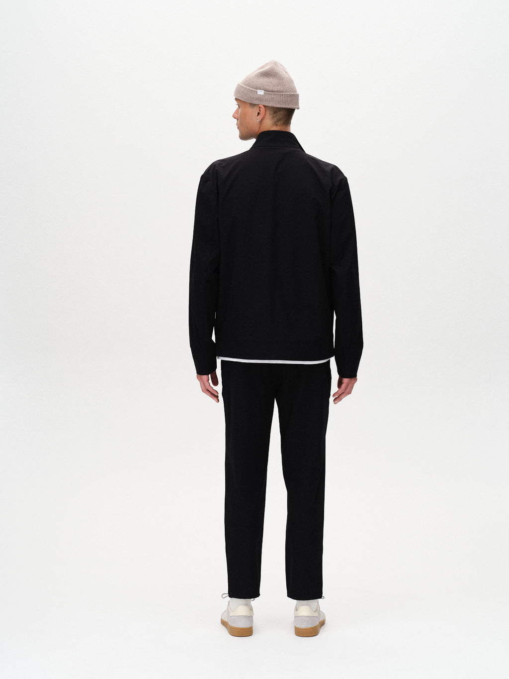 A person stands facing away in a Gabba Monza Tech - Black jacket, relaxed straight fit black pants with an elastic waist, white shirt, beige beanie, and white sneakers against a plain white background.