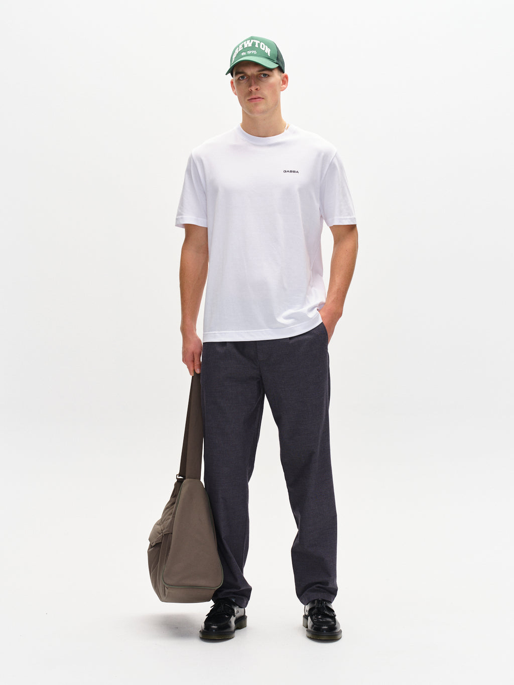 A person stands against a plain background wearing a white t-shirt, Gabbas Napoli 19 Carre pants in navy known for their versatility, and black shoes. They have a green cap and are holding a large beige bag at their side, exuding an effortless comfort.