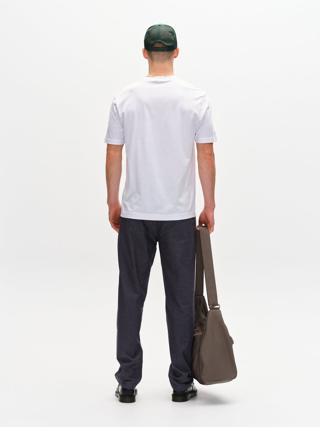 A person wearing a white T-shirt, Gabba Napoli 19 Carre pants in navy, and a green cap stands facing away, holding a large brown bag. The plain white background emphasizes the outfits versatile design that effortlessly combines comfort and style.