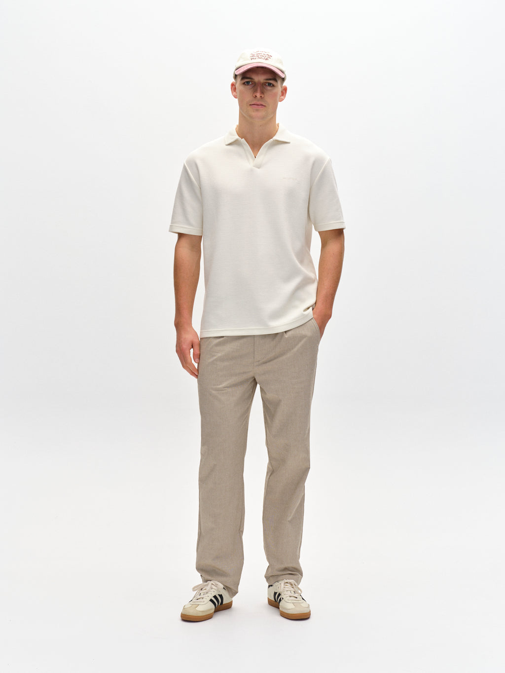 With a white polo shirt, Gabbas Napoli 19 Carre pants in sand, and a white cap against a plain background, they exude classic style. Sporting white sneakers with black accents and one hand in their pocket, they effortlessly blend comfort and sophistication.