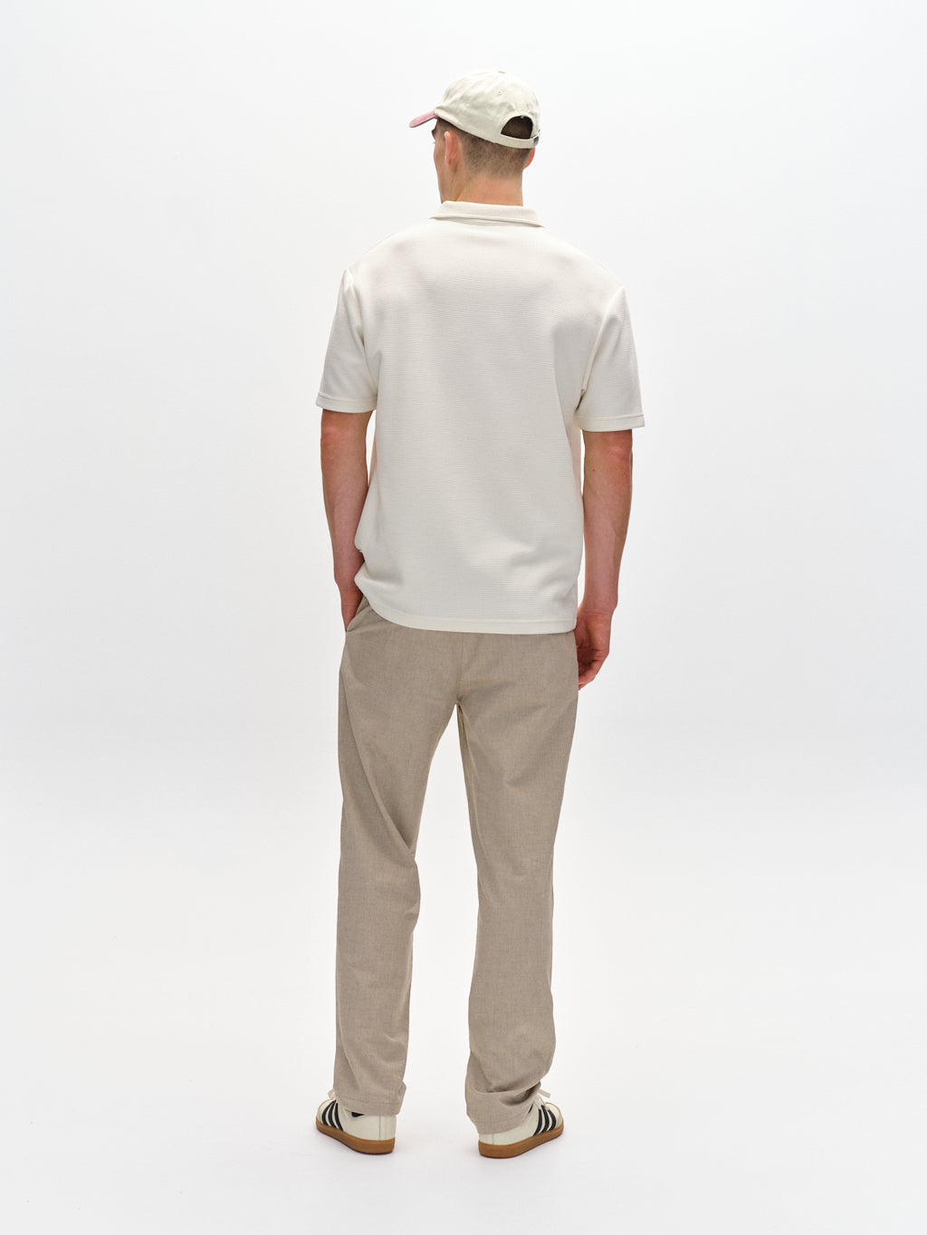 Wearing the Gabba Napoli 19 Carre pants in Sand, paired with a beige cap and white polo shirt, this individual showcases classic style. Standing against a plain white background, their striped sneakers add a touch of comfort to the ensemble.