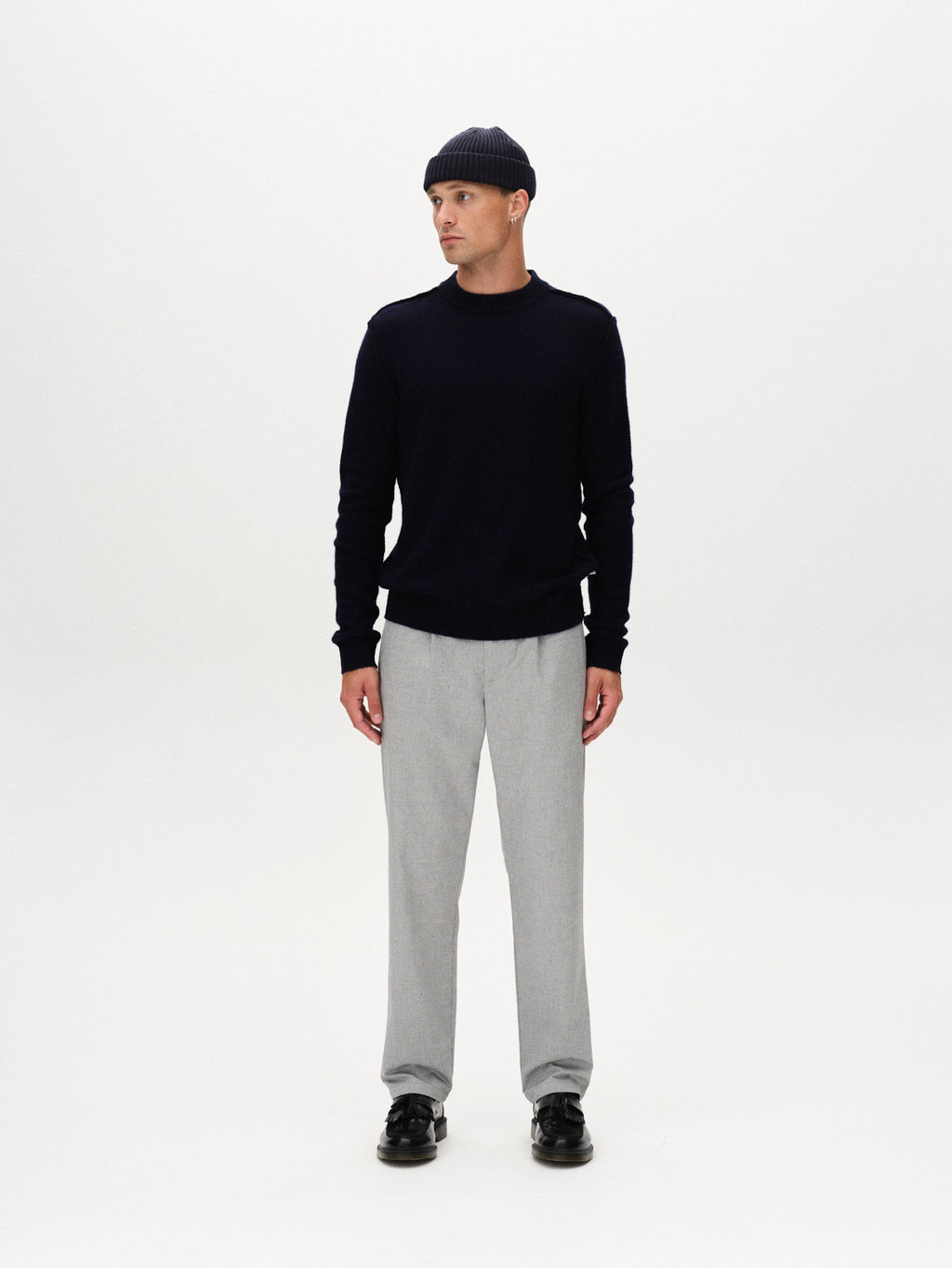 A person in a navy beanie, navy sweater, Gabba Napoli 19 Carre - Lt. Grey pants, and black shoes stands against a plain white background, looking slightly to the side.