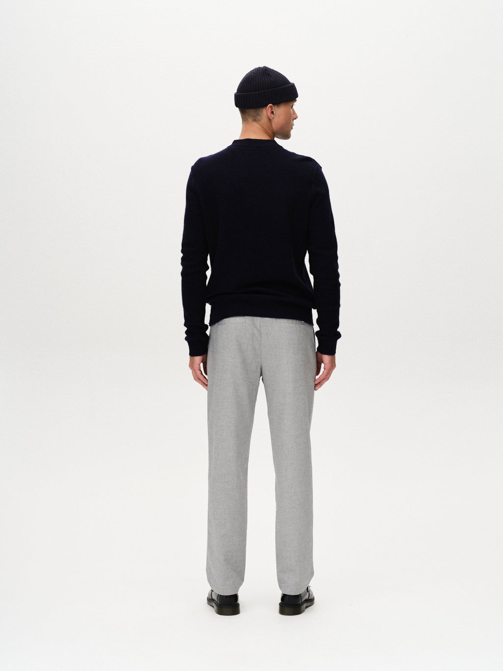 Against a plain white background, a person faces away from the camera wearing a navy sweater, black beanie, black shoes, and Gabba’s Napoli 19 Carre - Lt. Grey pants, highlighting a timeless and classic style.