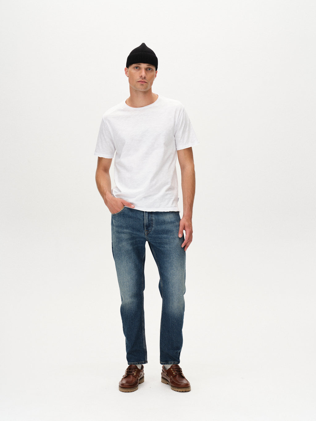 A man stands against a white background, wearing the Gabba Kostant New SS - White relaxed fit t-shirt, paired with a black beanie, blue jeans, and brown boat shoes, one hand in his pocket and a neutral expression.