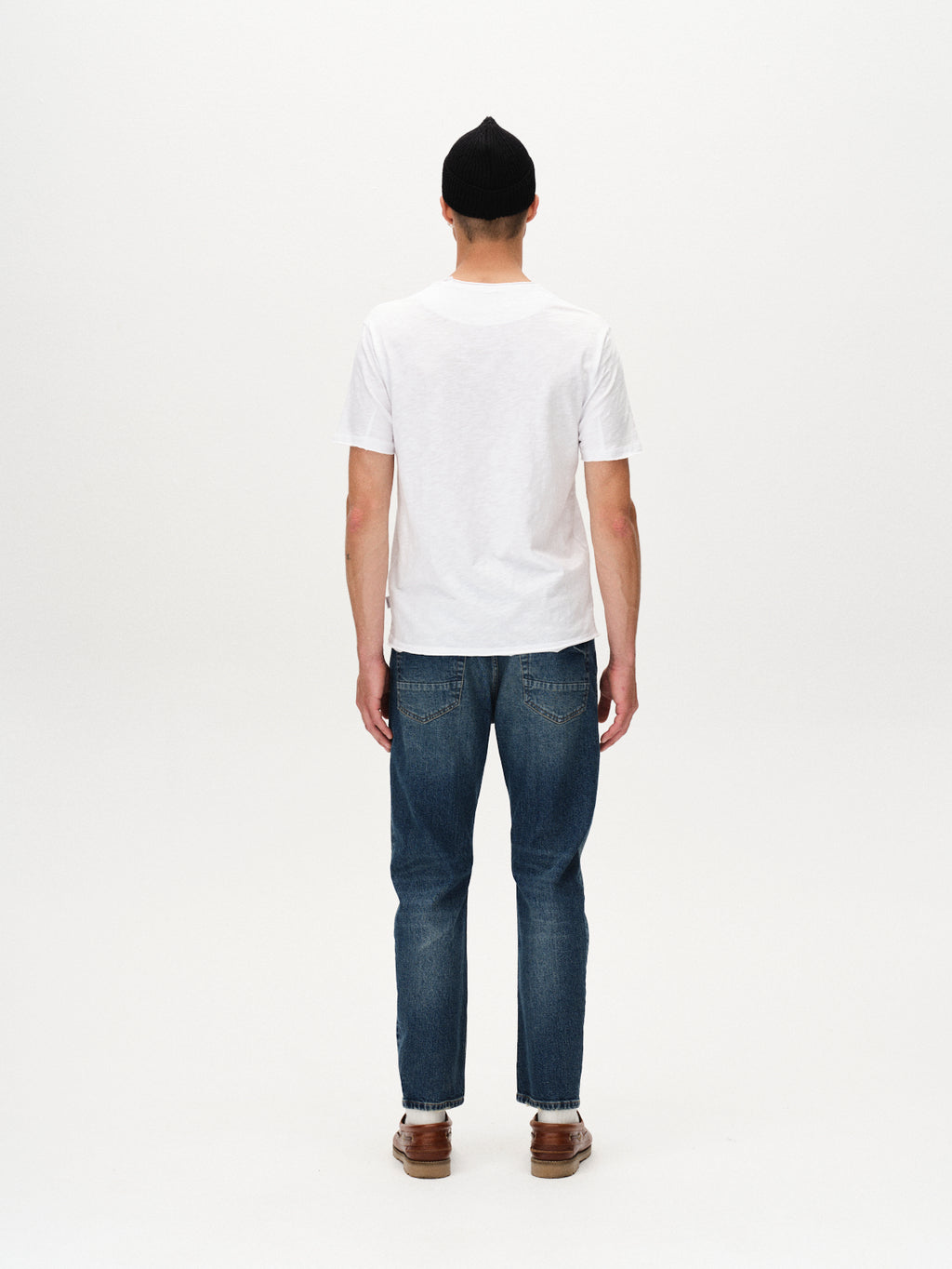 A person stands facing away from the camera against a plain white background, wearing the Gabba Kostant New SS - White t-shirt, a black beanie, blue jeans, and brown shoes.