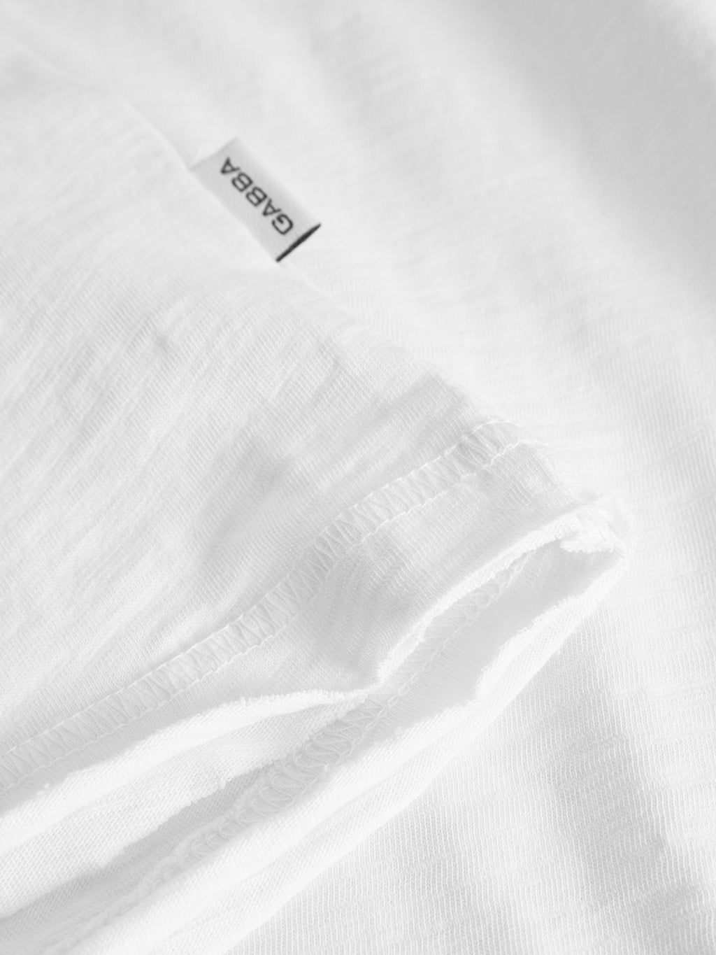 Close-up of the Gabba Kostant New SS t-shirt in white, made from lightweight slub fabric, featuring a folded sleeve and a visible white tag with “GABBA” in black text.
