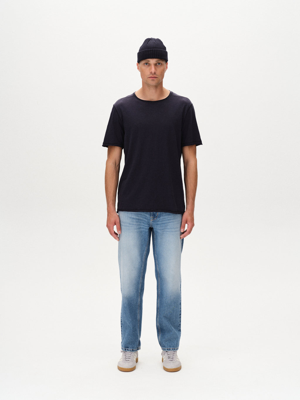 A person stands facing forward against a plain white background, wearing the Gabba Kostant New SS - Navy t-shirt, a dark beanie, light blue jeans, and light-colored sneakers.