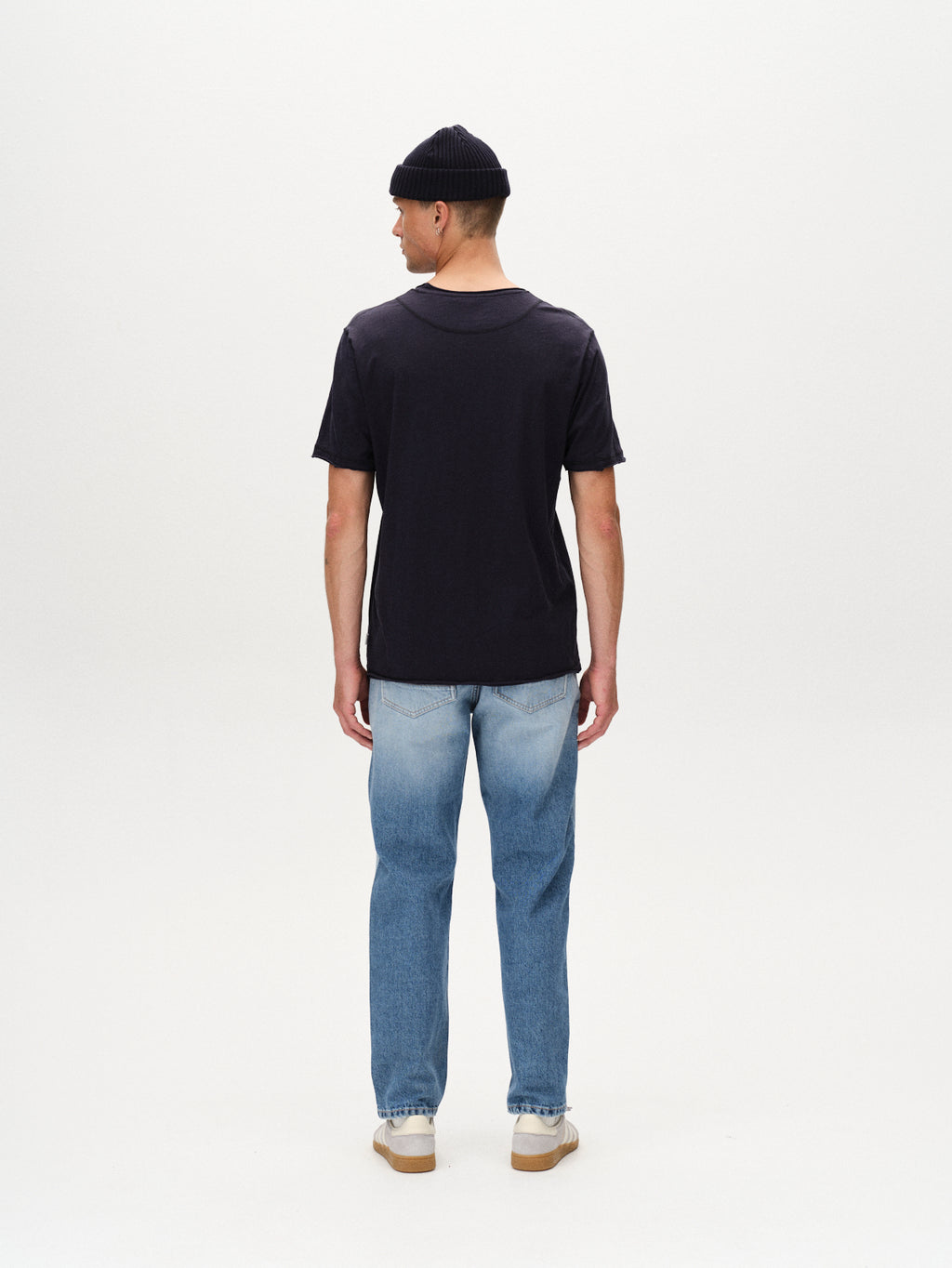 A man faces away from the camera, wearing the Gabba Kostant New SS navy t-shirt, a black beanie, light blue jeans, and white sneakers against a plain white background.