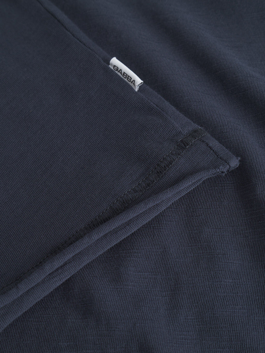A close-up of the Gabba Kostant New SS shirt in navy shows its textured slub fabric, visible flatlock stitching, and a small white GABBA tag attached to the seam.