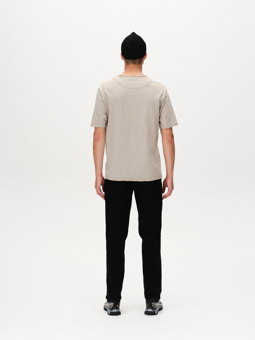 A person faces away from the camera against a plain white background, wearing Gabbas Kostant New SS - Grey Paloma shirt, black pants, black beanie, and sneakers.