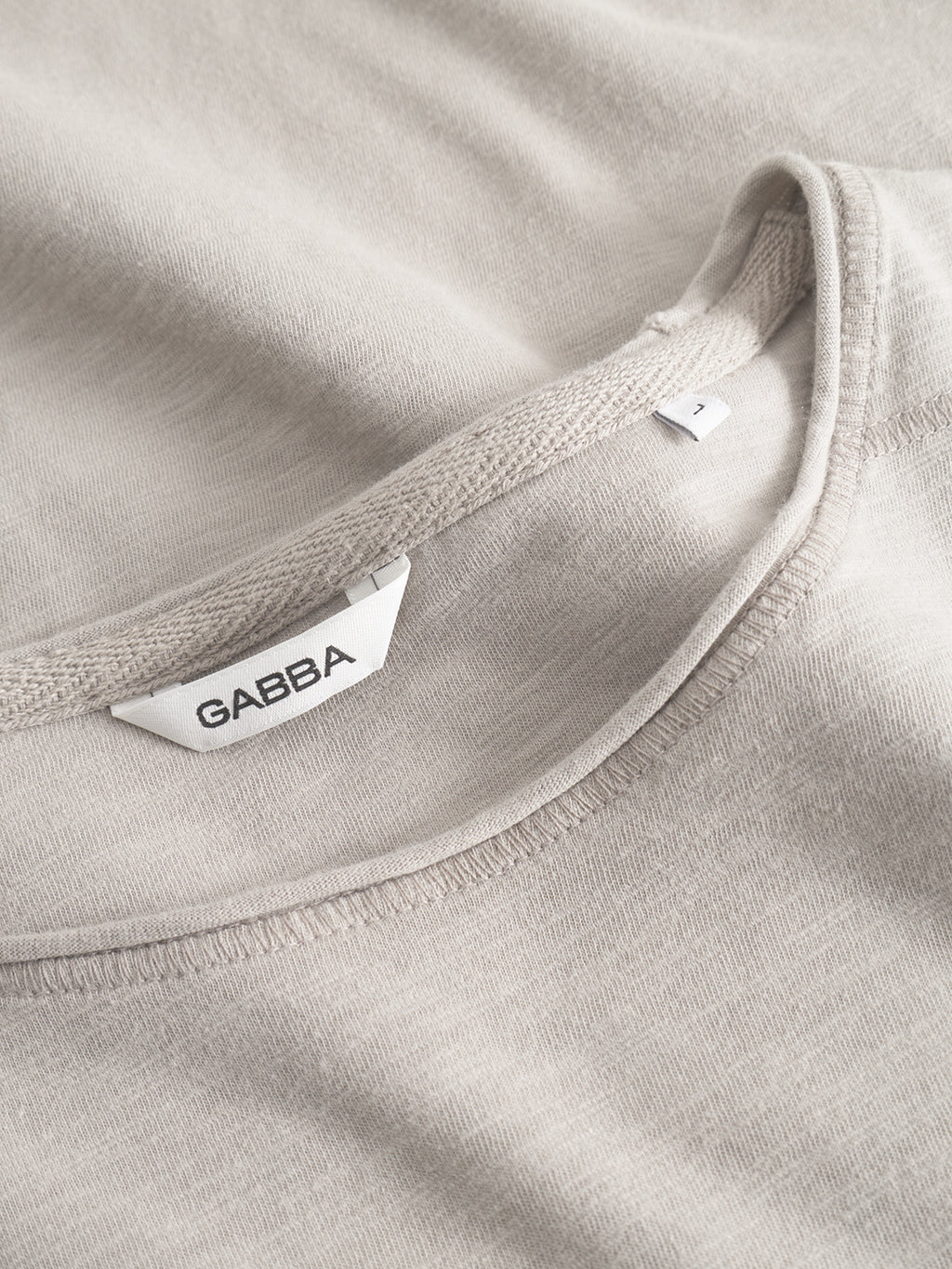 Close-up of the Kostant New SS - Grey Paloma T-shirt by Gabba, highlighting the neckline with a soft slub fabric and precise flatlock stitching. The inner label displays GABBA in black letters.