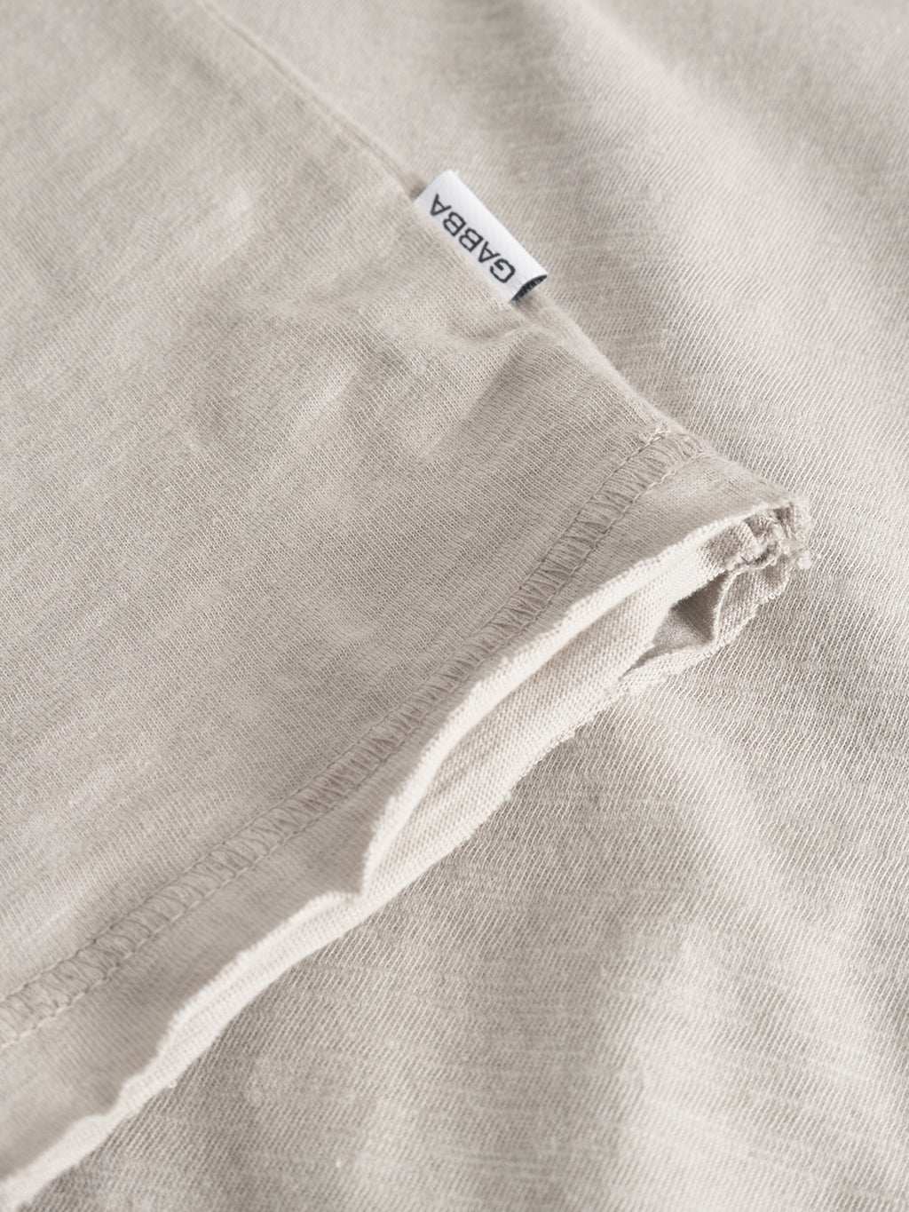 Close-up of the Kostant New SS - Grey Paloma T-shirt by Gabba, showing light beige slub fabric, detailed flatlock stitching, and a small white Gabba tag.