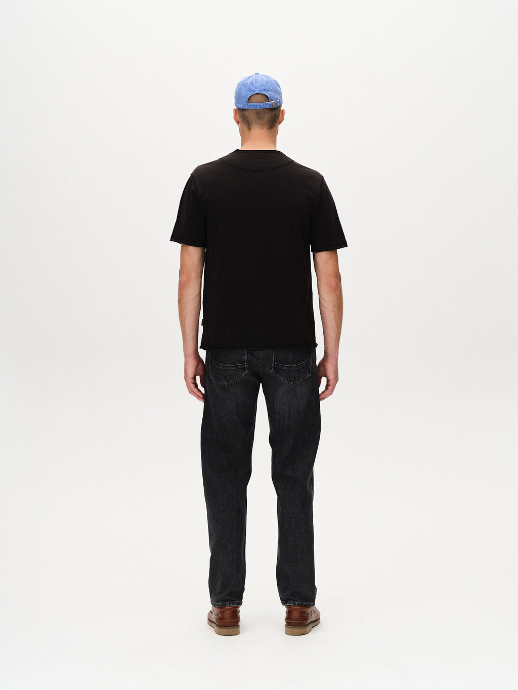 A man faces away from the camera, wearing the Gabba Kostant New SS - Meteorite relaxed fit black T-shirt, a light blue cap, dark jeans, and brown shoes against a plain white background.