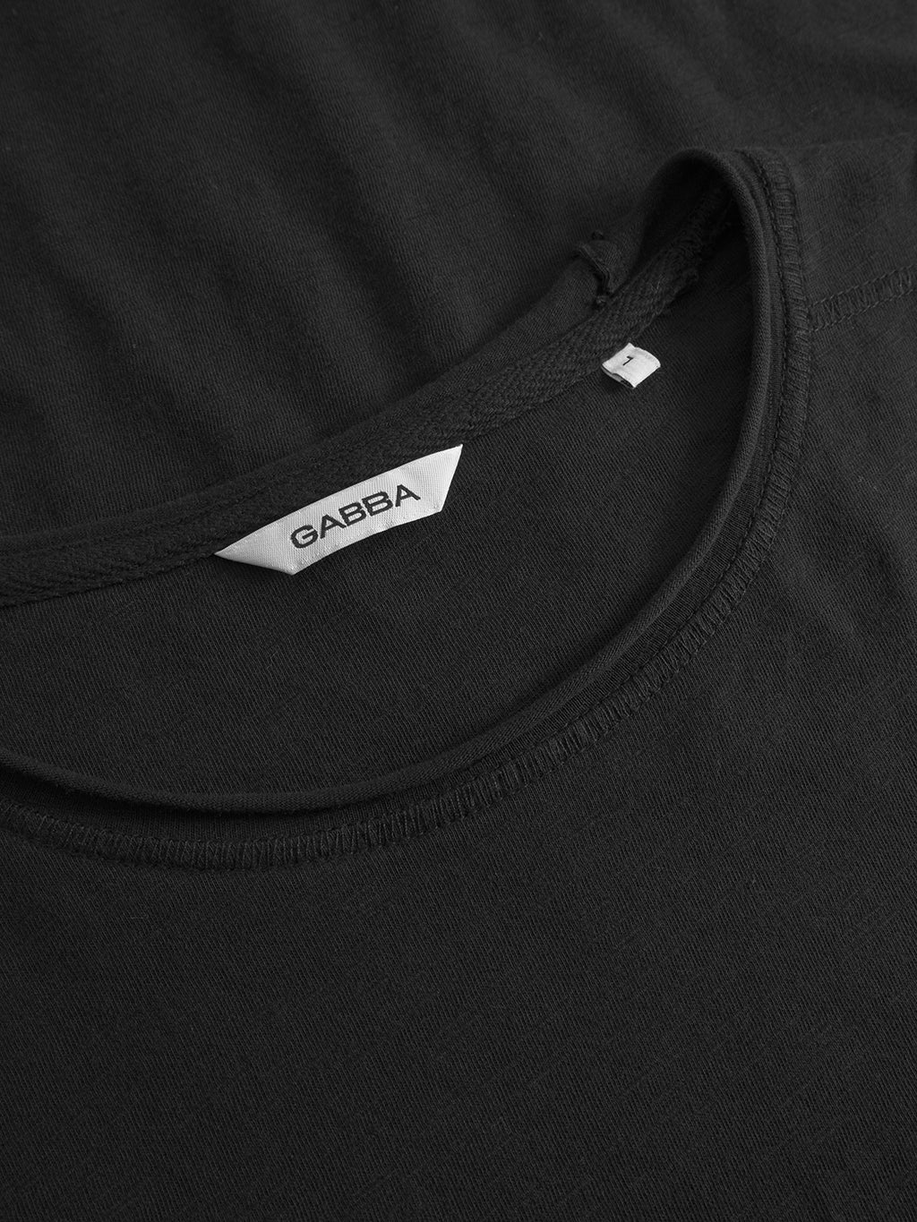Close-up of Gabbas Kostant New SS - Meteorite, a black crew neck T-shirt in slub fabric, with a white Gabba label sewn at the neckline, set against a matching black background.