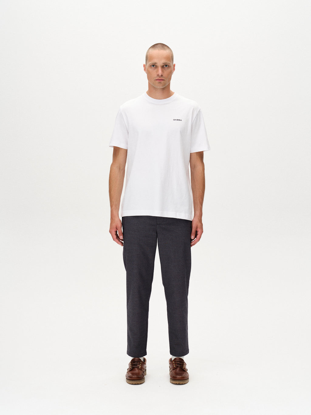 A man with short hair and a neutral expression stands facing forward against a plain white background, wearing the Gabba Monza Button Carre shirt in navy, relaxed straight pants with an elastic waist in dark gray, and brown shoes.