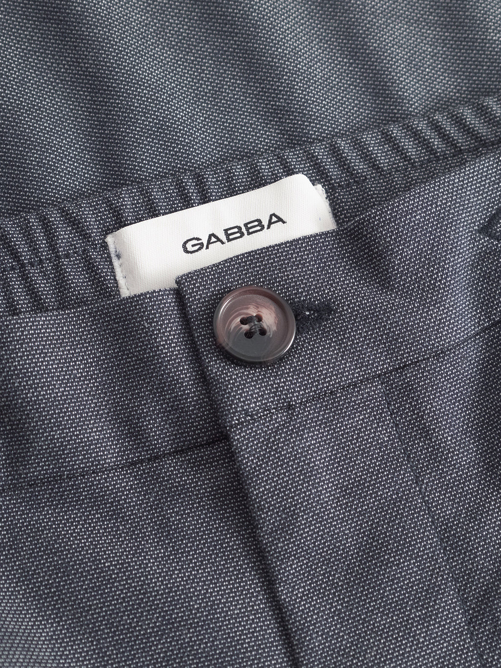 Close-up of a pair of relaxed straight pants with a visible white Gabba label above the waistband. The gray textured pants feature a Monza Button Carre from the navy collection, secured through a buttonhole.