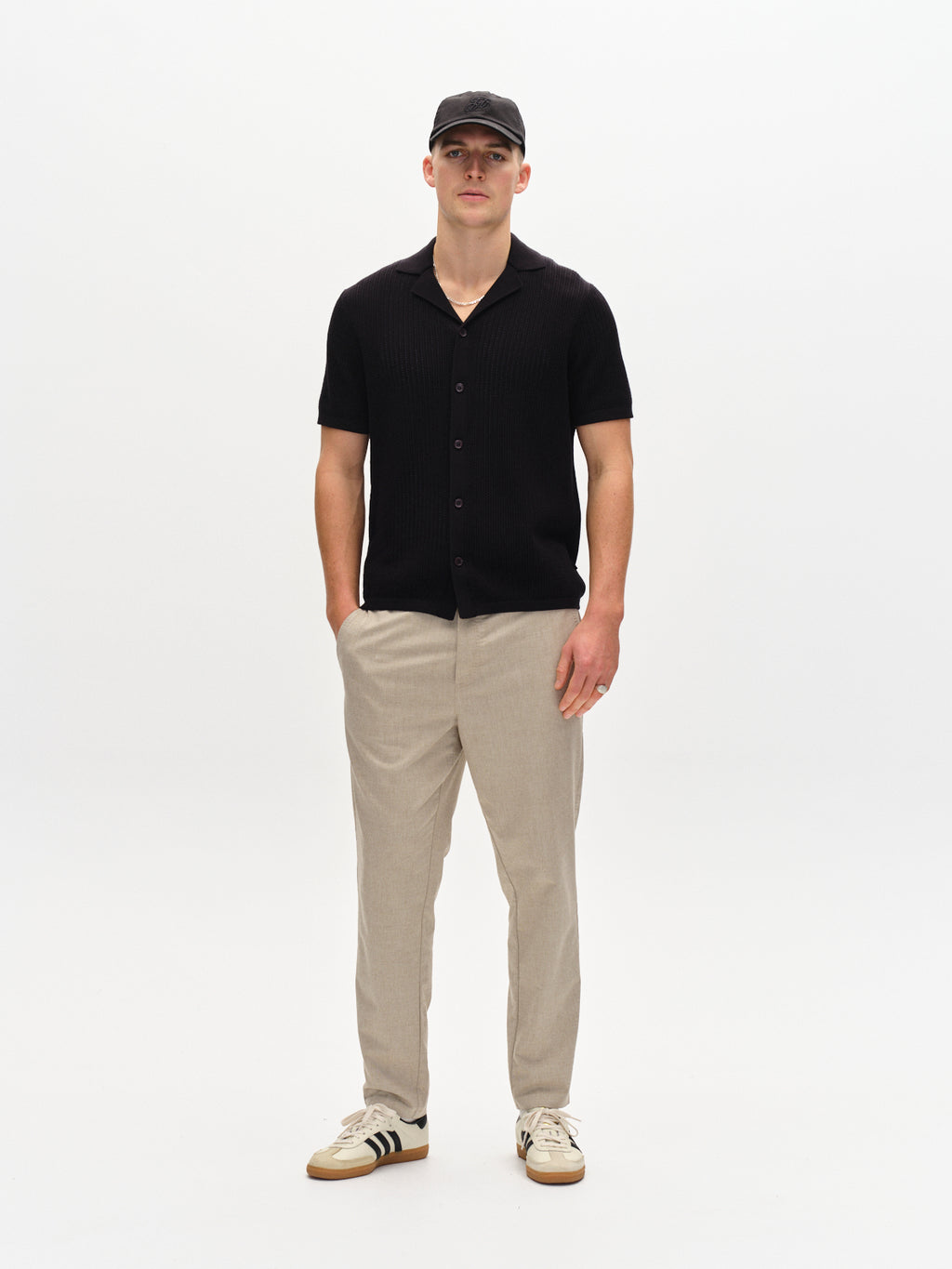 A person stands against a plain background wearing a sand-colored Gabba Monza Button Carre short-sleeve shirt, light-colored relaxed straight pants with an elastic waist, a black cap, and white sneakers with black stripes. Hands in pockets, they maintain a neutral expression.