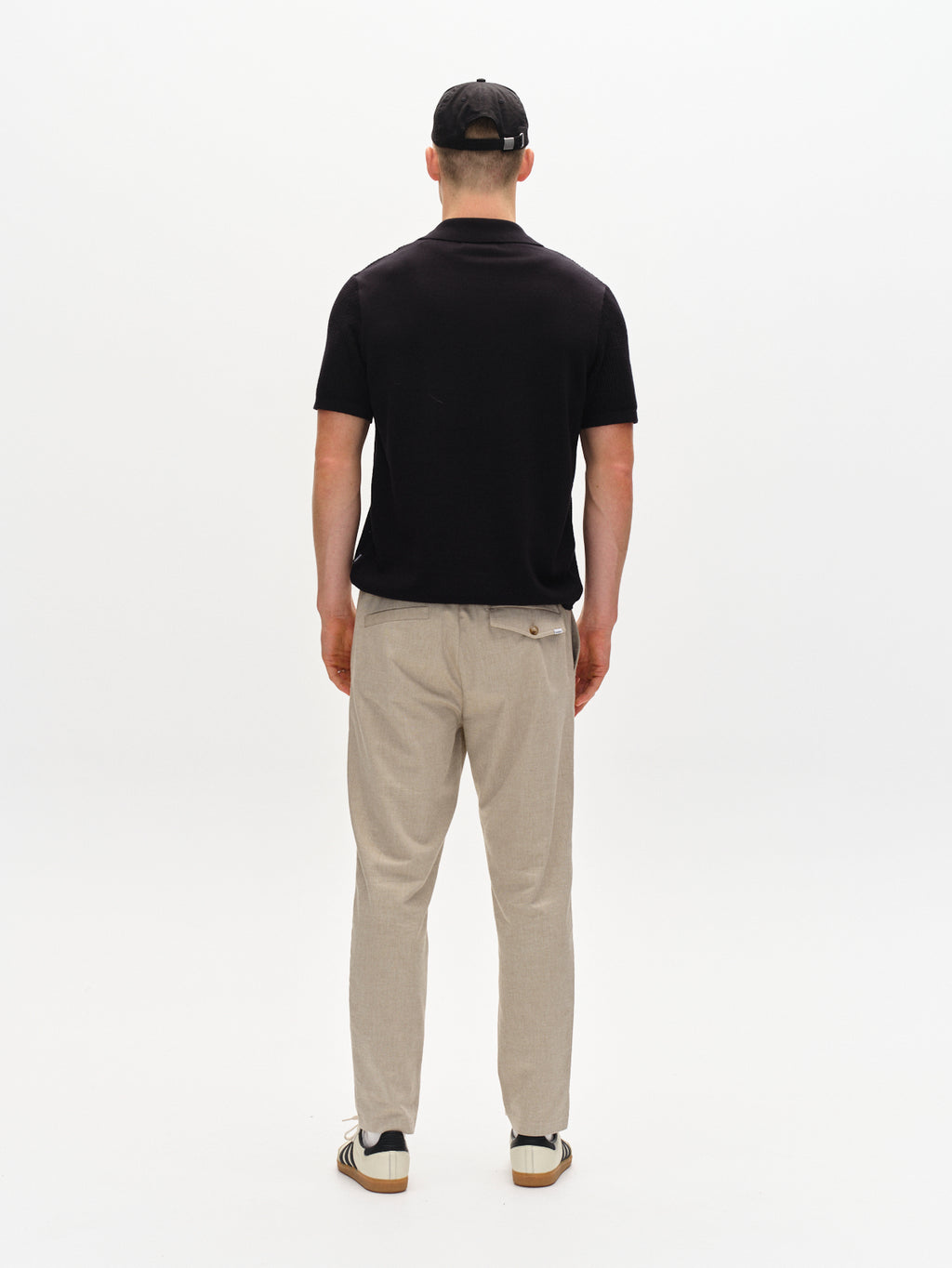 A person stands with their back to the camera against a white background, wearing a black t-shirt, Gabbas Monza Button Carre in sand—relaxed straight pants with an elastic waist—a black baseball cap, and black and white sneakers.