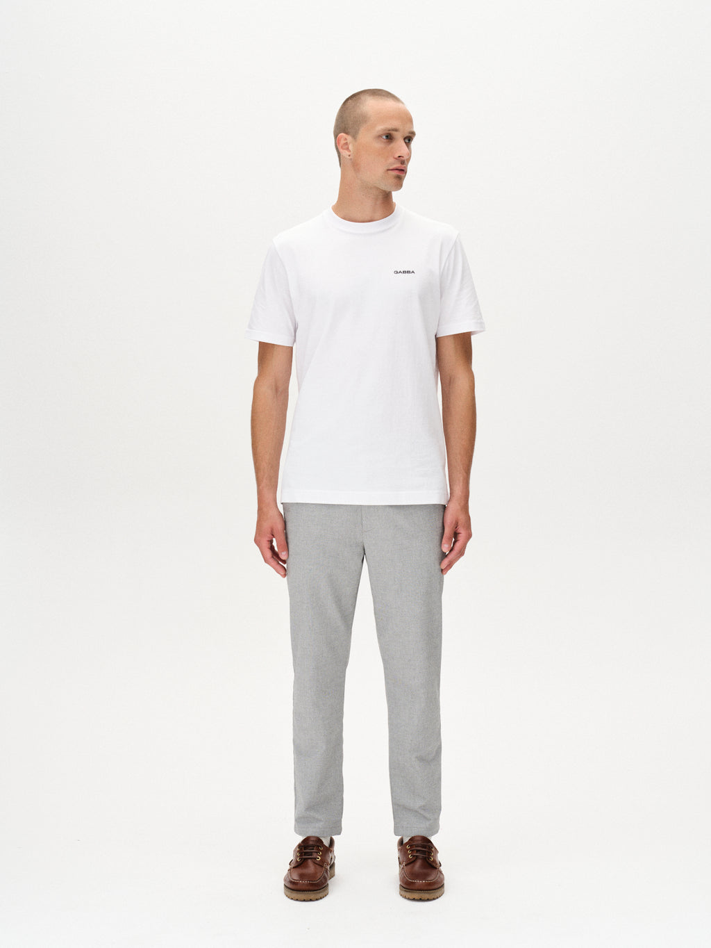 A man stands at an angle against a plain white background, wearing Gabbas Monza Button Carre pants in light grey, paired with a simple white t-shirt and brown boat shoes.