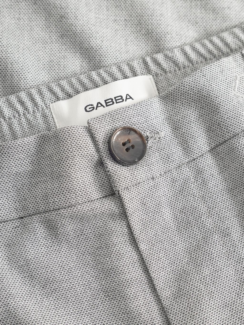 Close-up of Monza Button Carre pants in light grey by Gabba, showcasing textured fabric, button closure, elastic waist for comfort, and a white Gabba label sewn inside—ideal for versatile styling.