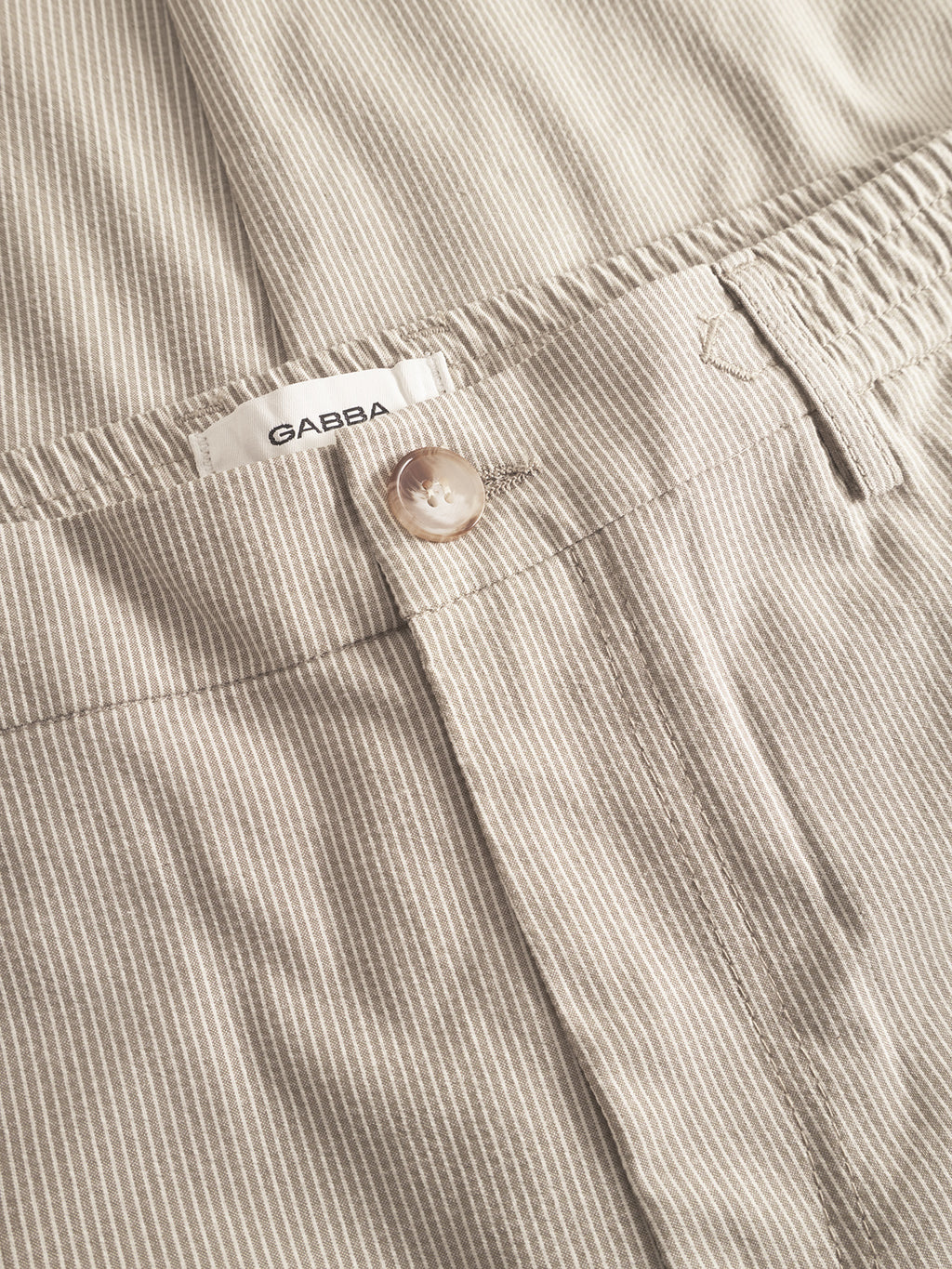 Close-up of Gabba Monza Button Com2 Cropped pants in khaki, featuring light beige pinstripes, elastic waist for comfort and style, a button, and a white Gabba label. The lightweight fabric has a finely textured appearance.