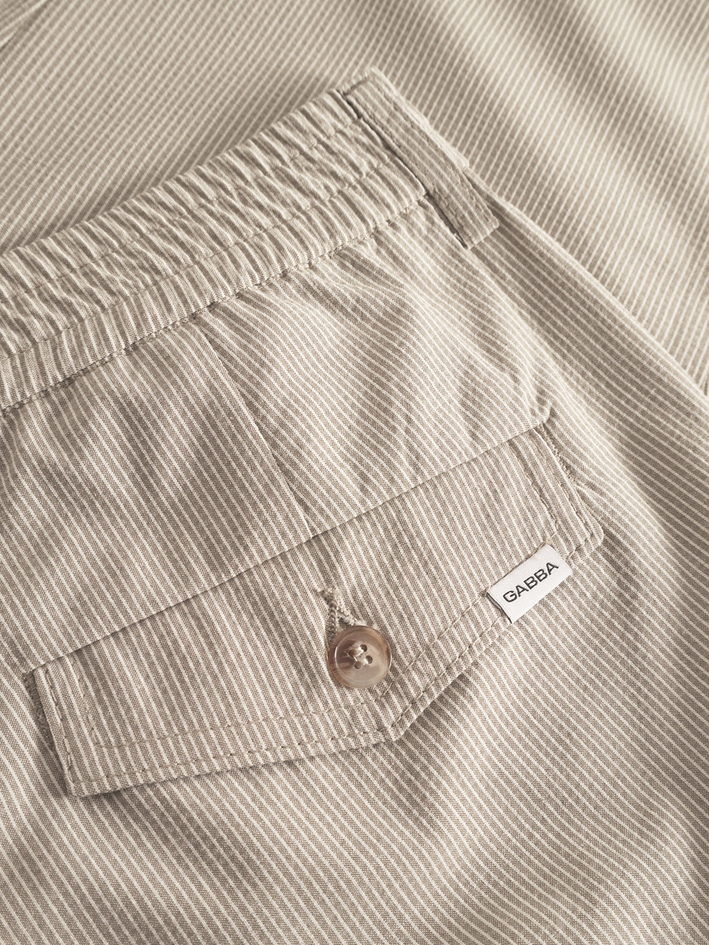Close-up of the Monza Button Com2 Cropped - Khaki by Gabba, showing beige and white vertical stripes, an elastic waistband for comfort and a stylish buttoned pocket with a small white Gabba label.