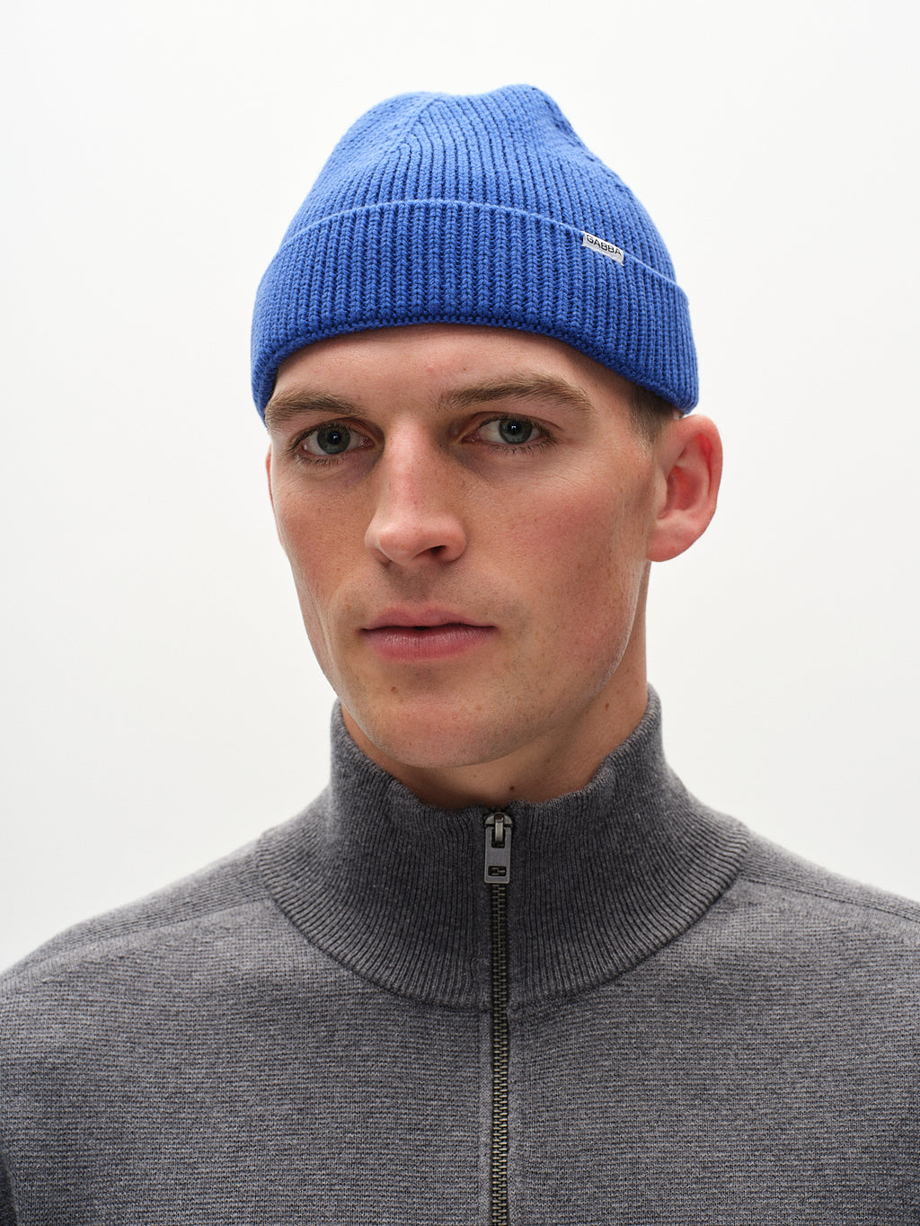 Wearing the vibrant blue New Mount Beanie by Gabba, with its distinctive small label, and a complementary gray high-collar zip-up sweater, a person gazes off to the side against a plain backdrop.