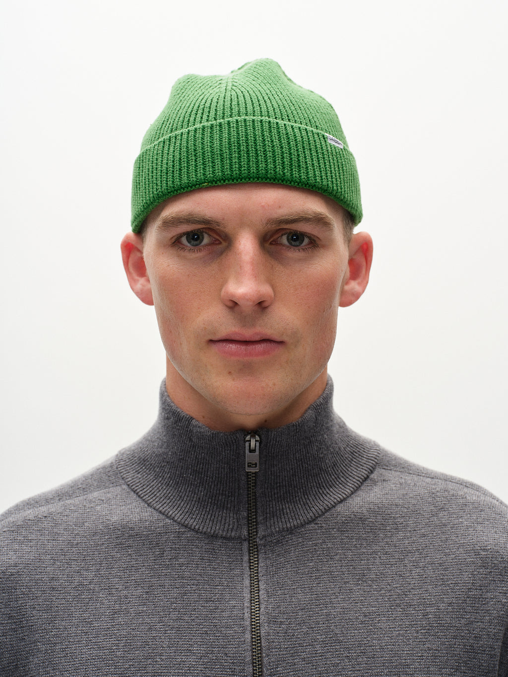 A person in a green New Mount Beanie by Gabba, featuring a ribbed knit design, and a gray zip-up sweater is centered against a plain light background. Their head and shoulders are shown with a neutral expression.