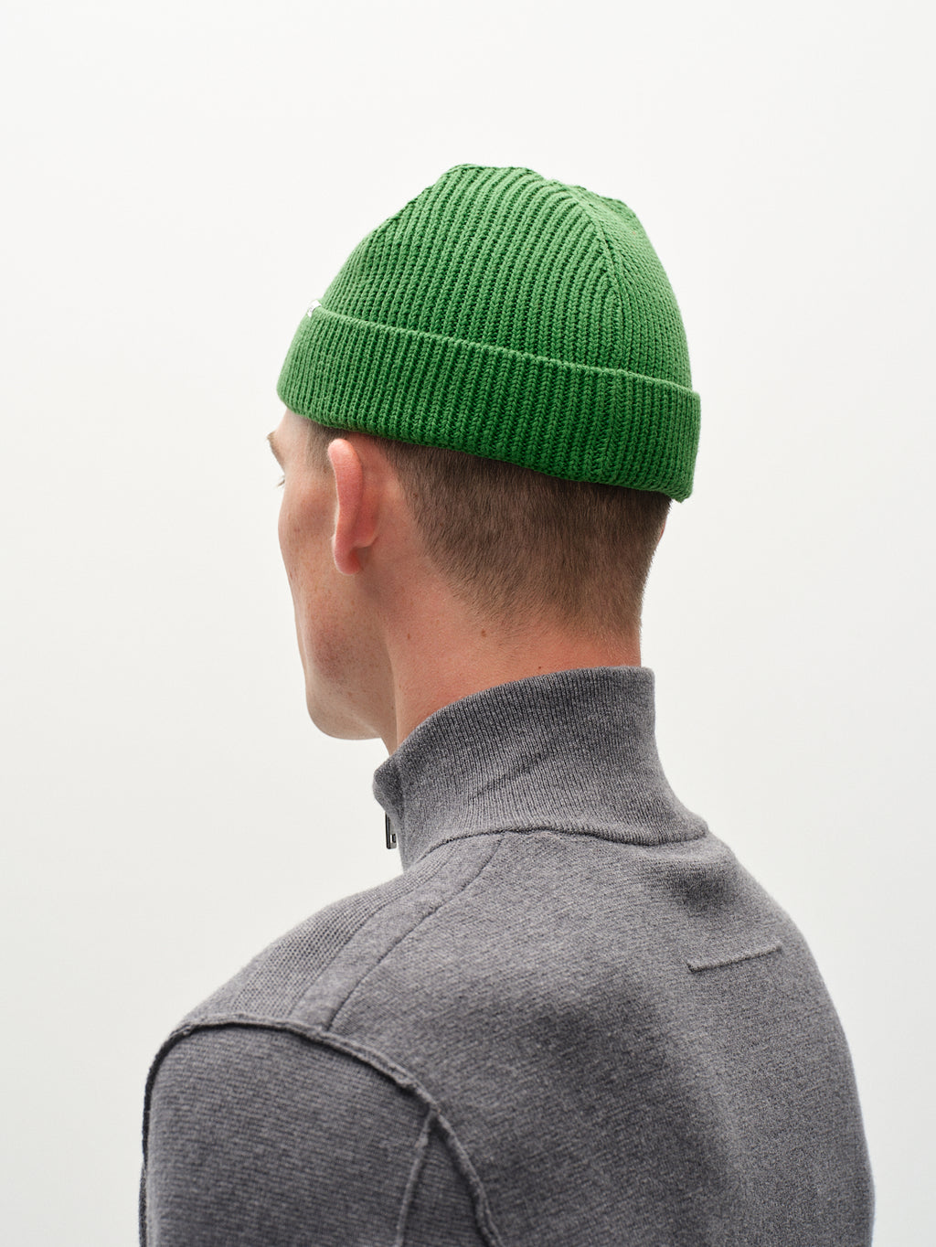 A person in a gray zip-up sweater is seen from behind wearing the New Mount Beanie - Green by Gabba, featuring a ribbed knit design. The vibrant color of the beanie stands out against the plain, light-colored background.