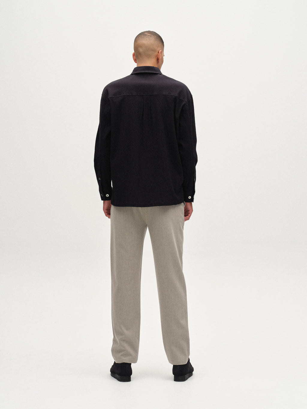 A person, facing away from the camera, wears a black long-sleeve shirt with Gabba's GABnapoli 19 Phin GRS - Sand straight-leg trousers and black shoes, set against a plain white background.