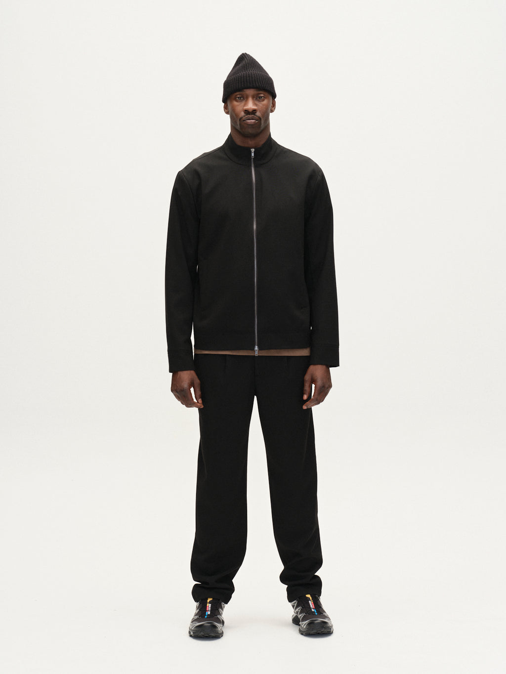A man stands facing forward against a plain white background, wearing Gabba Napoli 19 Phin GRS - Black trousers, paired with a black beanie, zip-up jacket, and sneakers. His arms are relaxed at his sides.