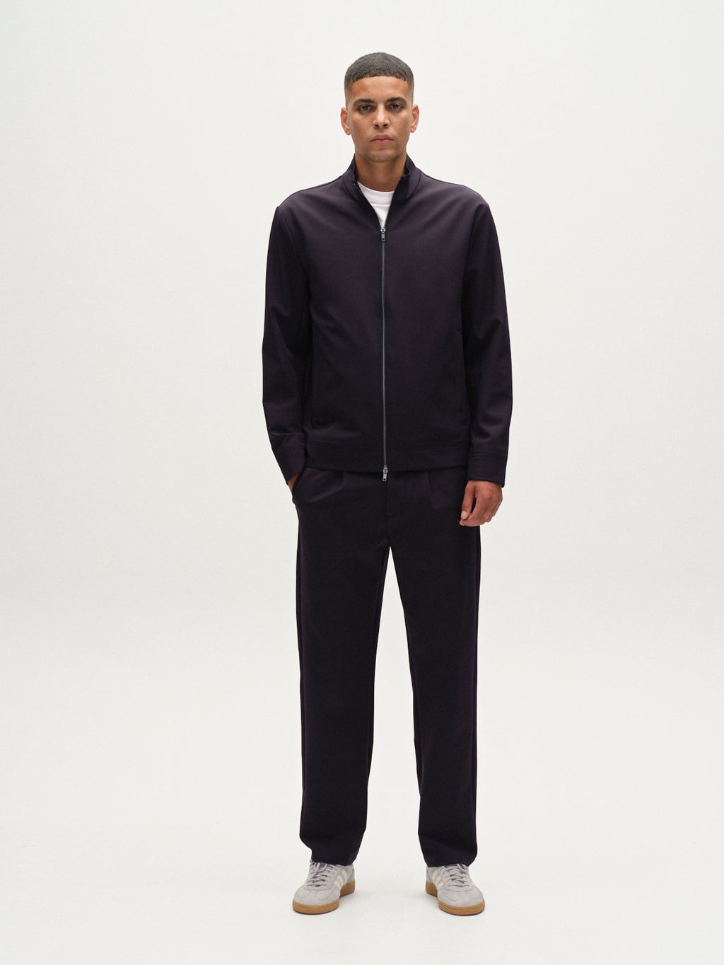 A man stands facing forward against a plain white background, wearing Gabba’s Napoli 19 Bascu - Navy zip-up jacket, wrinkle-resistant trousers, a white t-shirt, and white sneakers. His hands are relaxed by his sides.