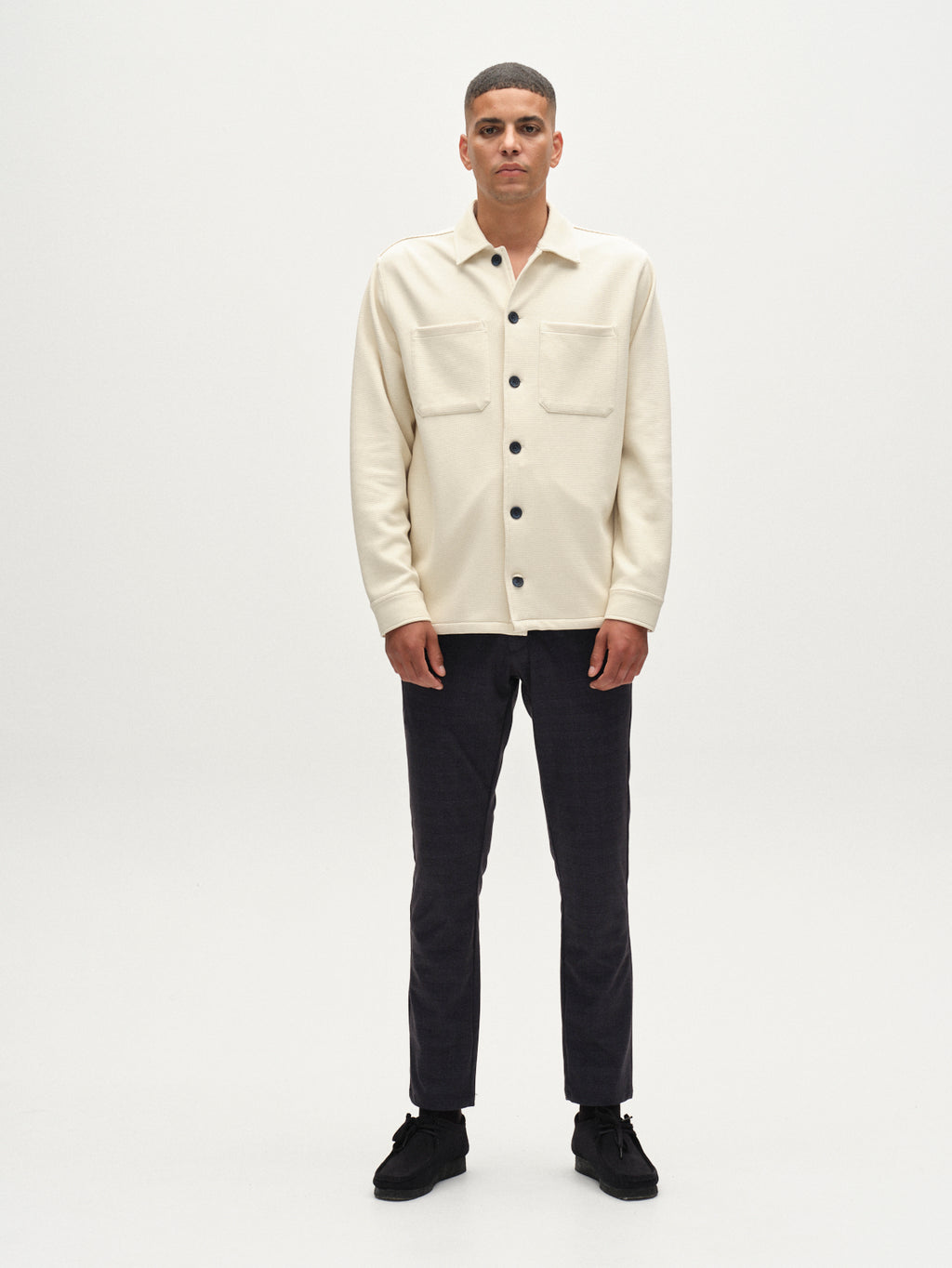 A man faces forward against a white background, wearing Gabba’s GABpaul Nygo – Navy slim straight trousers, a cream button-up shirt with front pockets, and black shoes.