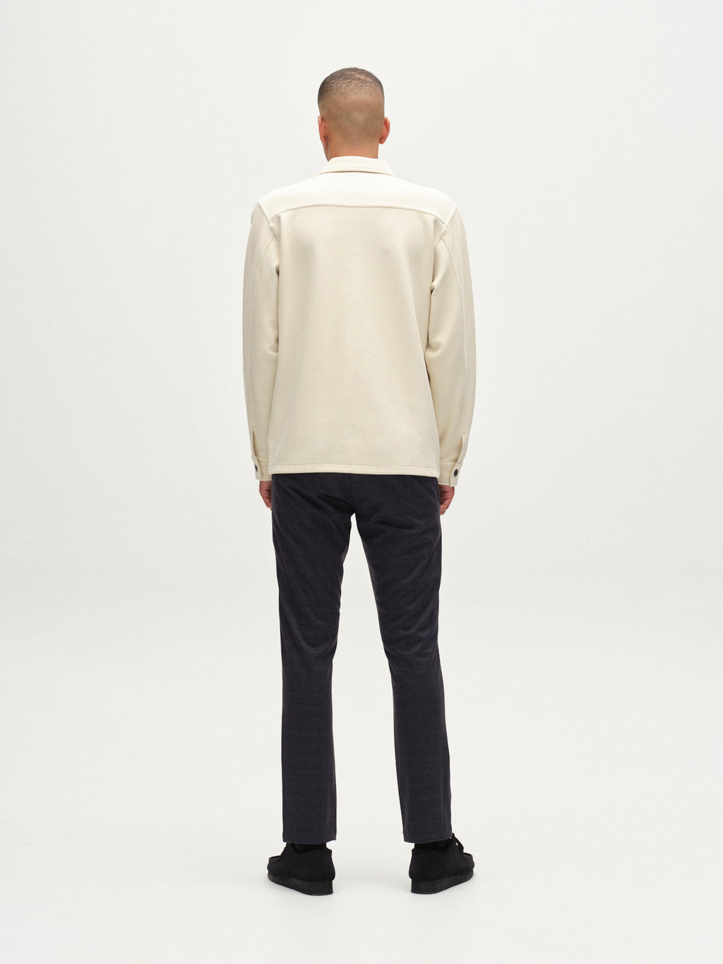 A person stands facing away from the camera, wearing a cream long-sleeve shirt, Gabba GABpaul Nygo navy slim straight trousers, and black shoes against a plain light background.