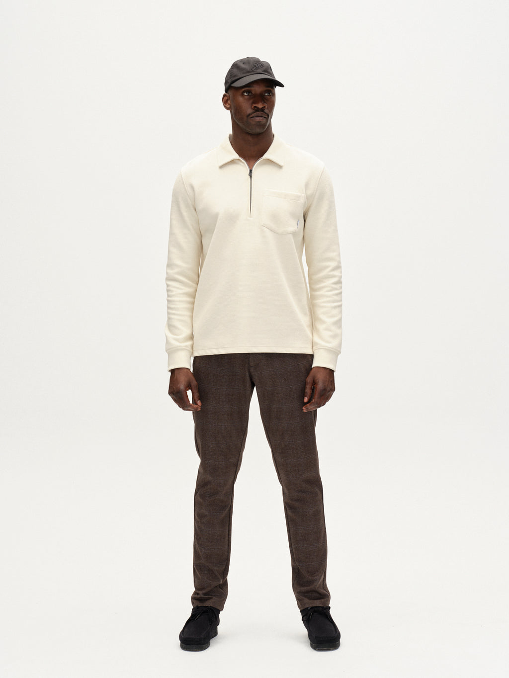 A man stands against a white background, wearing a cream zip-neck shirt, Gabba Paul Nygo dark brown slim straight stretch trousers, black shoes, and a dark baseball cap.