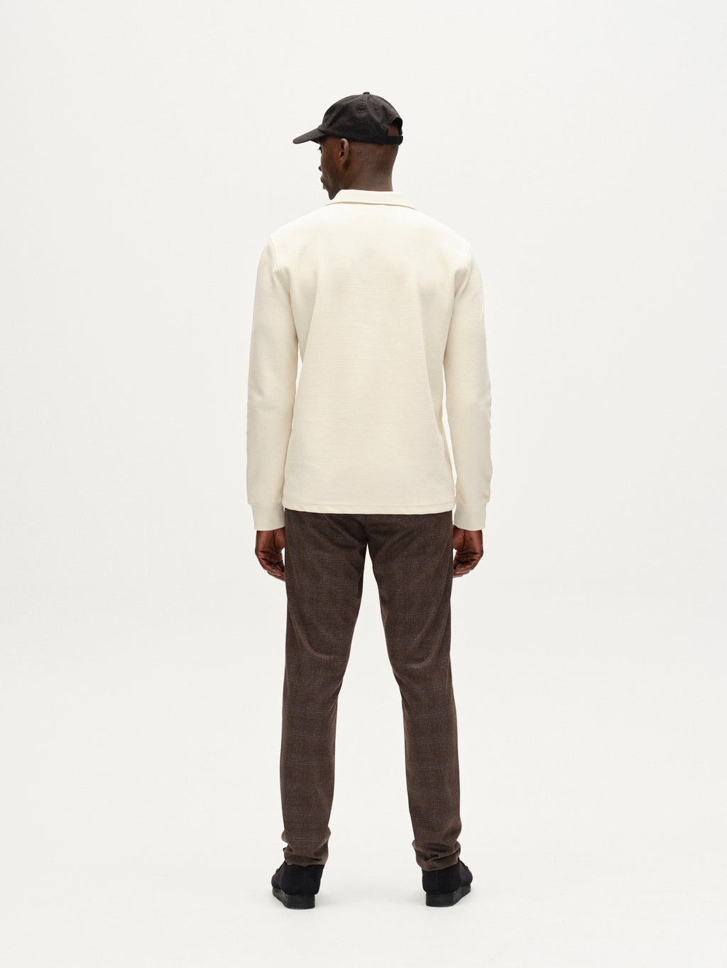 A man, seen from behind, wears a cream long-sleeve shirt, black shoes, a dark baseball cap, and Gabba Paul Nygo - Dark Brown slim straight trousers with stretch comfort, set against a plain white background.