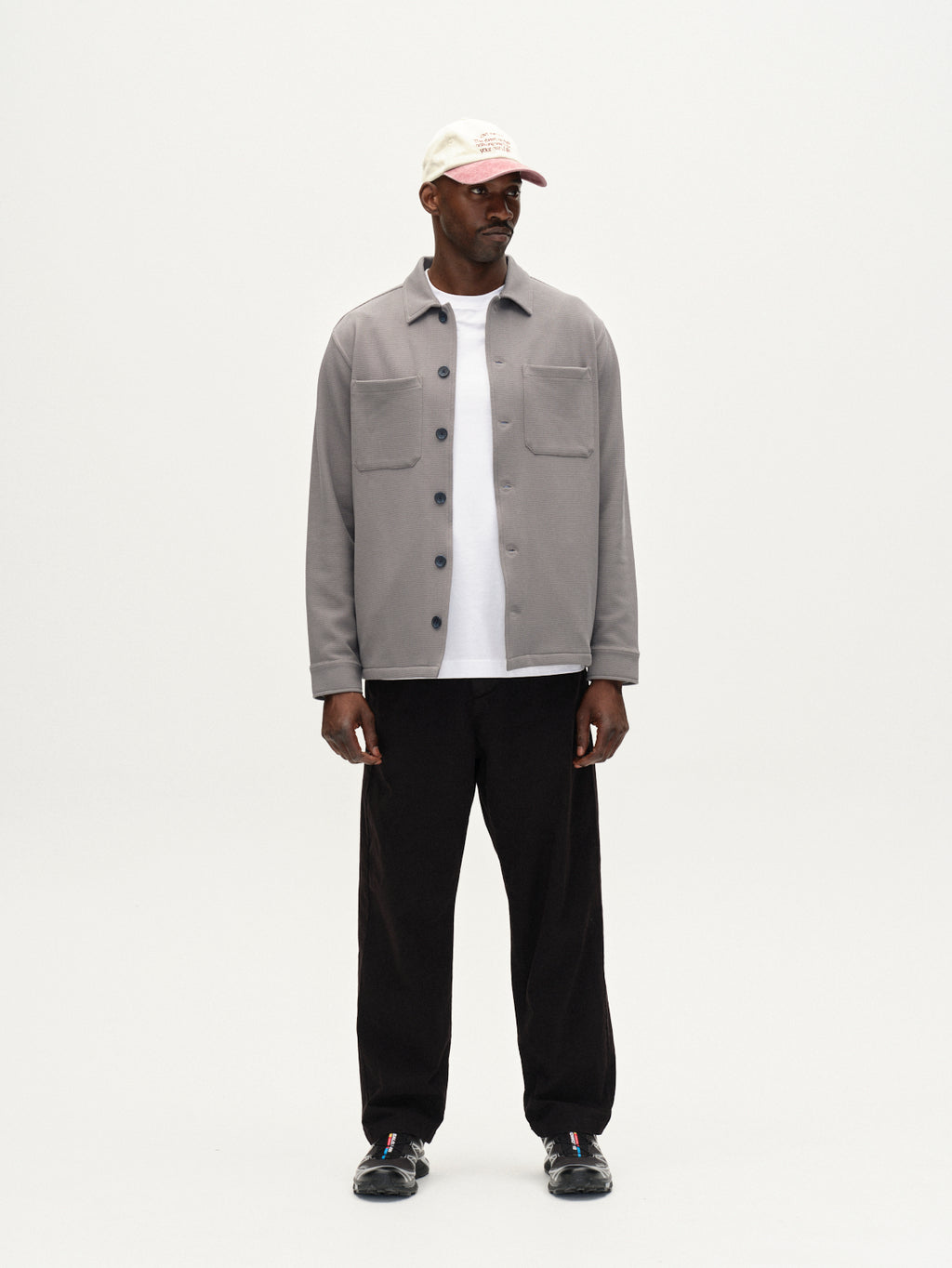 A man wears the Gabba Como Waff overshirt in Dust Sage Green over a white t-shirt, paired with black pants, black shoes, and a white cap with red text, standing against a plain white background.