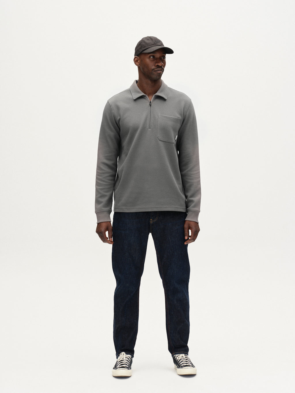 A man stands against a plain white background, wearing the Gabba Island Waff long-sleeve shirt in Dust Sage Green, paired with dark blue jeans, black sneakers with white laces, and a gray cap.