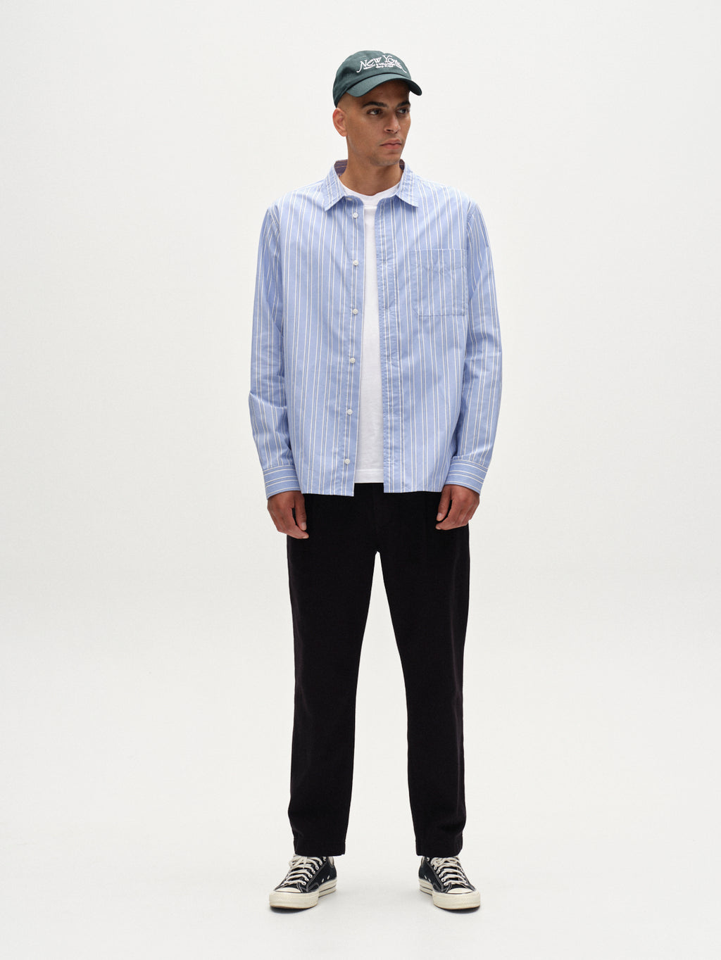 A man stands against a plain white background wearing a green baseball cap, a light blue and white striped button-up over a white tee, relaxed-fit trousers, and Gabba’s Joe-Pas Vill - Black sneakers.