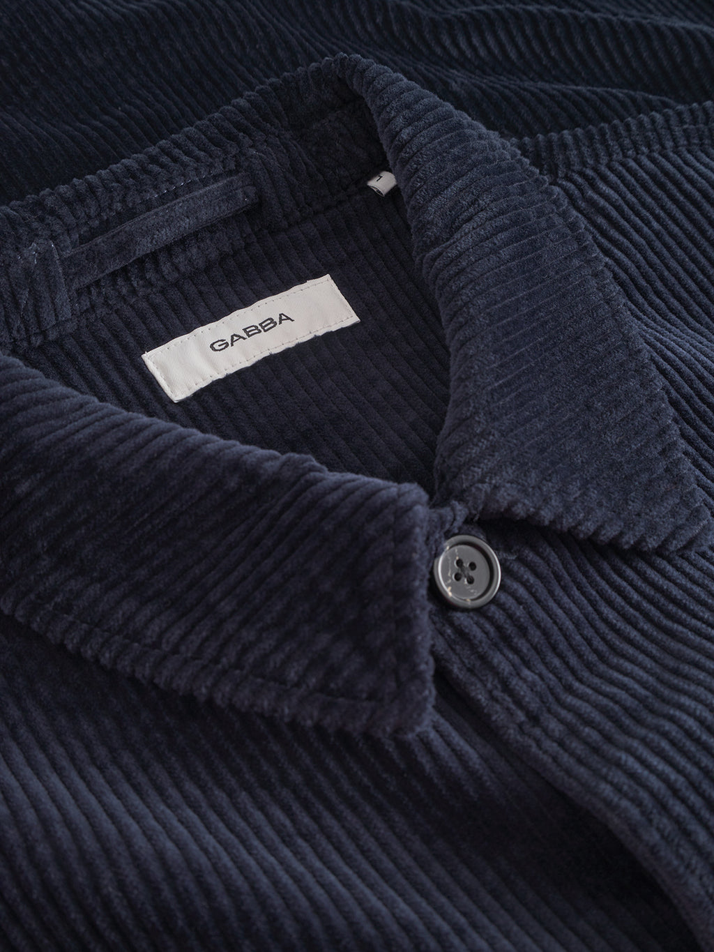 Close-up of the Libre AX Cord - Navy shirt by Gabba, featuring navy blue brushed stretch corduroy, a visible collar, black button, and a white label with the Gabba brand name inside the neckline.