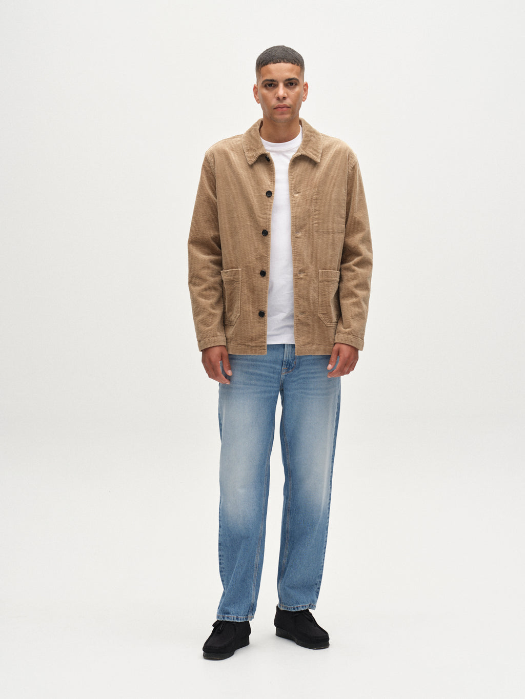 A man stands facing forward against a plain white background, wearing the Gabba Libre AX Cord shirt in sand over a white t-shirt, paired with light blue jeans and black shoes.