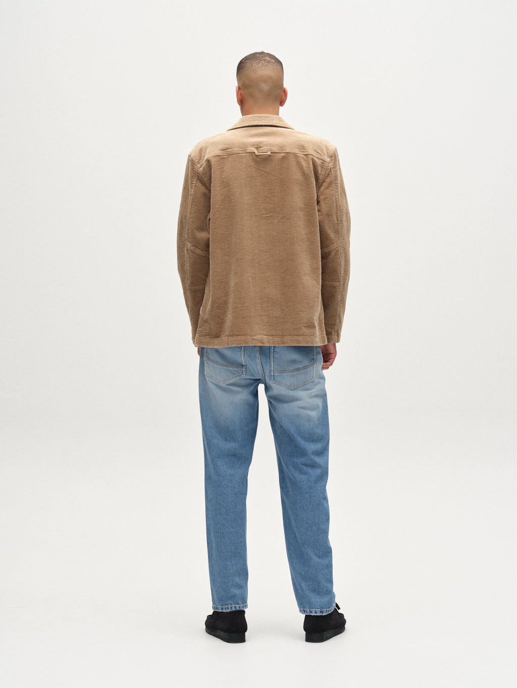 A person stands facing away from the camera, wearing the Gabba Libre AX Cord shirt in sand, light blue jeans, and black shoes against a plain white background.